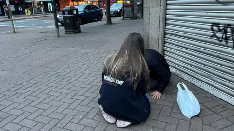 A woman with long highlighted hair kneels down to speak to someone on the street in Belfast. She is wearing a black Welcome Organisation fleece.