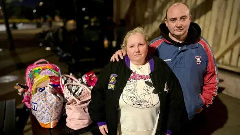 A man and woman wearing a jumper and large coats look at the camera. In the background, their belongings are in a B&M bag, a pink handbag and a children's backpack