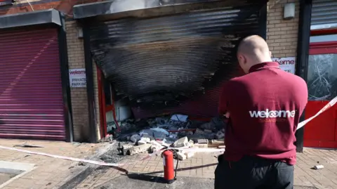 Pacemaker The exterior of the Welcome Organisation building with a burned garage door which is warped as a worker in a maroon polo shirt surveys the damage