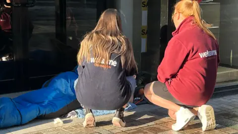 BBC Welcome Organisation workers kneel on the ground in an attempt to speak to a person in a blue sleeping bag