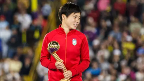 Getty Images Choe Il-son, wearing his country's red tracksuit, smiles as she holds the Golden Ball trophy