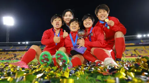 Getty Images North Korea players celebrate and show off their medals to the camera. They're sitting on the turf, which covered in gold-colured tickertape.