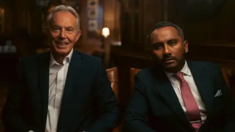 BBC Tony Blair and Amol Rajan are sat in a dimly lit room. They are both wearing suits and looking directly at the camera.