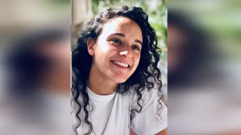 Mandy Damari  Emily Damari, a young woman with long dark, curly hair, smiles while sitting in the sunshine, wearing a white T-shirt 