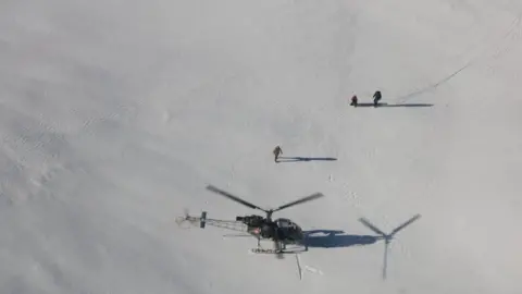 Reuters An aerial shot of a helicopter having landed on snow near two people with a third person walking between them