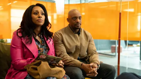 Curfew / Paramount+  Alexandra Burke wears a magenta pink jacket and is sat on a sofa next to Oliver Alvin-Wilson who is wearing a brown coat. They are both staring into the distance.