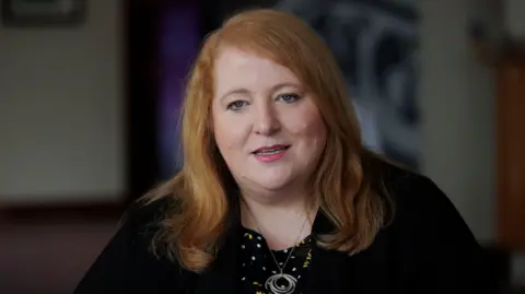 Liam McBurney/PA Wire Justice Minister Naomi Long looking to camera, wearing a black jacket, a black, white and yellow top with polka dots and floral patterns, and a necklace with concentric rings. 