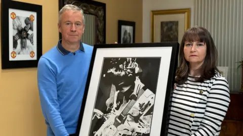 Peter Dolan and his wife Niamh Dolan standing together and looking to camera. Between them they are holding a larger black and white photo of their late son Enda playing guitar.