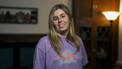 A young woman in a purple t-shirt looks at the camera