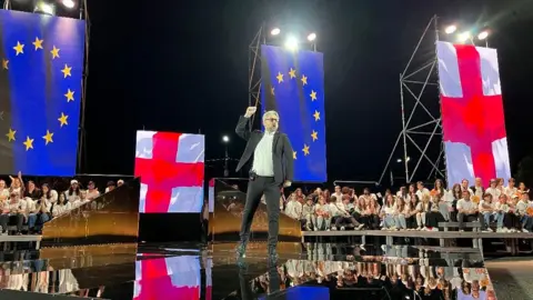 Nika Gvaramia stands on stage pumping his fist in the air, with audience members seated around. The stage is backlit by European Union and Georgian flags