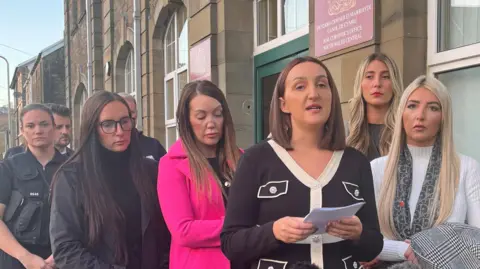 five women stand together with one, Amy Smith, holding a paper and speaking 