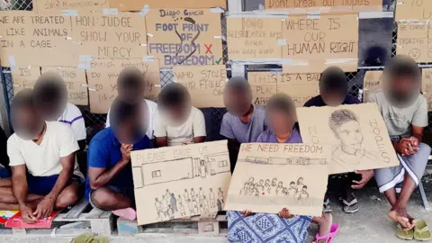 BBC Tamil migrants on Diego Garcia, with their faces blurred, holding protest signs reading "please save us" and "we need freedom"with other signs attached to a fence behind them