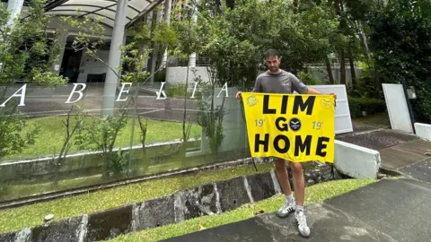 Dani Cuesta/X A man outside a residential apartment complex holding a yellow banner that reads LIM GO HOME