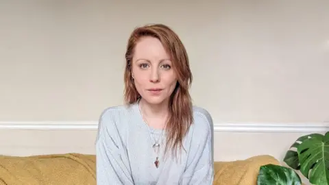 Hannah Clarke Hannah Clarke, with long ginger hair, sitting on a sofa and wearing a grey top with a plant seen next to her