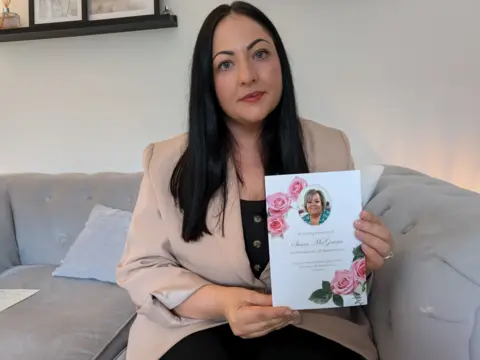 Jade Campbell, who has long dark hair, sits on her grey couch at home looking into the camera with a neutral expression. She is holding a funeral programme for her aunt. She is wearing a peach blazer with the sleeves rolled up, a black top and black trousers.