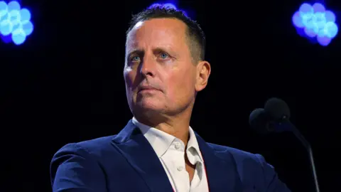 Reuters Richard Grenell, who has short brown hair, wears a blue suit jacket and white shirt, as she stands in front of a microphone onstage at a convention