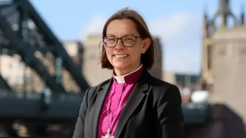 DIOCESE OF NEWCASTLE Helen-Ann Hartley wears a clerical collar with a pink shirt, black blazer, and silver cross chain, in a picture taken on the streets of Newcastle