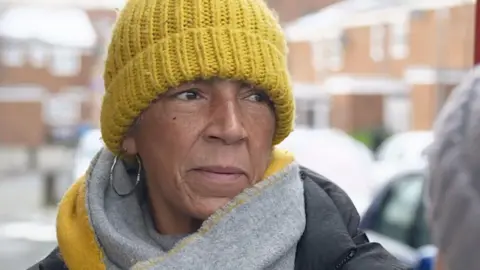 Angela stands in a street wearing a yellow hat and grey and yellow scarfand hoop earrings.