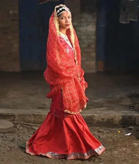 Innovation for Change A girl poses for the camera wearing colourful red clothing and jewellery, she's standing outside on a street and looks directly at the camera. The scene is well lit which makes the clothing looks vibrant. 