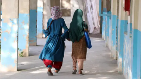 EPA Two Afghan girls walk down an outside corridor with blue and white walls, their hair covered. They wear brightly-coloured clothes, and the sun is shining