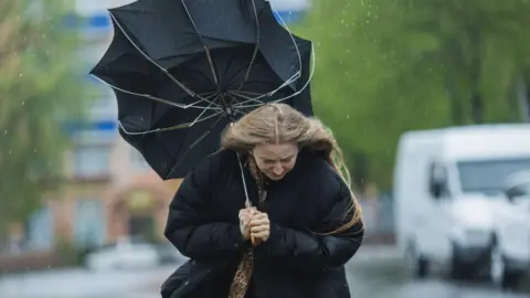 Getty Images A woman in a large black puffer coat is holding a black umbrella which is turned inside out. The woman is looking down as she is being rained on. In the background are blurred images of vehicles and a street.