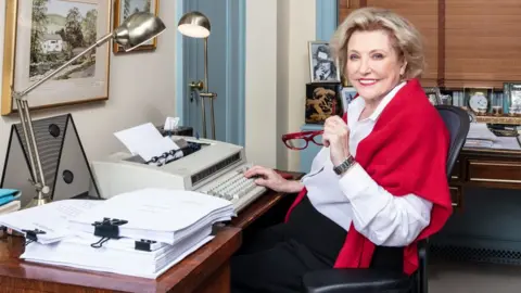 Caroll Taveras/Bradford Enterprises Barbara Taylor Bradford sitting at a typewriter in an office. she is wearing a white shirt and a red shawl and is holding her red glasses in her left hand.