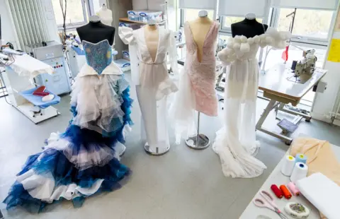 StoryShop Four dress designs are tacked onto mannequins in a work shop. The first one has a blue sparkly bodice with a blue and white mesh skirt. The second is white and has a deep V neck and asymmetric detailing in the shoulders and skirt. The second  is light pink and about knee length with a very deep V down to about the hips. The fourth has white balls about the size of a fist going diagonally across the dress. It has a short skirt with a longer mesh skirt over the top.