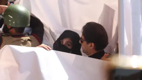 EPA Bushra Bibi and Imran Khan are shielded by a white sheet as they arrive at a courthouse. Bibi has turned to look at the camera, her hair is covered so is her nose and mouth.