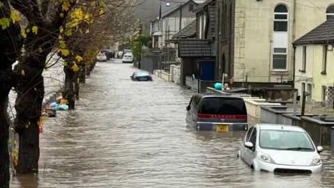 BBC Cars stranded in flooded street as man uses a bucket to tip water over wall