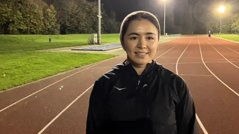 Shariqua Ahmed/BBC Fatima Painda wearing a black tracksuit jacket, standing on an athletics track 