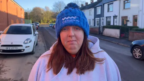 BBC Melissa McComb - a woman stands in the middle of a street where terraced houses are behind her and looks at the camera. She has medium-length brown hair and is wearing a blue beanie had with 'Wild Atlantic Way' embroidered in white and a grey hooded jumper.