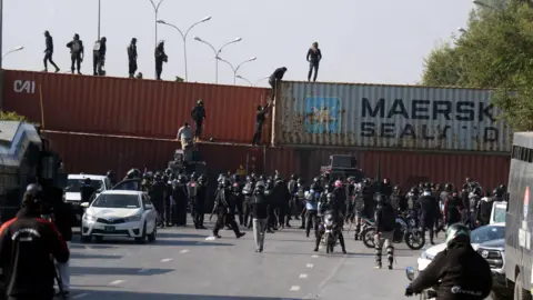 EPA A multi-lane highway with four shipping containers in the middle of it. People are climbing over the containers while police watch on