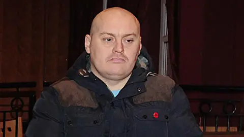 Pacemaker Ian Ogle is seen wearing a navy coat with a poppy pin.
He is bald, with a light stubble and frowning.
