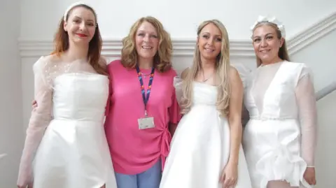 StoryShop Three woman (models called Ilona Chmiela, Reanna McGuire and Mandy Gibson) in white bridal dresses stand arm in arm. Another woman, second from the left, is casually dressed in a pink top and blue trousers. They are standing in what looks like a stairway with a white wall in the background.