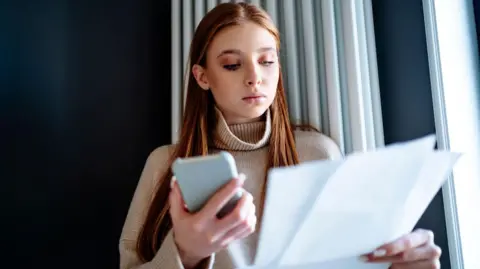 Getty Images Young woman stands in front of a radiator holding a smartphone and a paper bill.