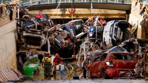 Reuters At the entrance to a road tunnel, two firefighters operate a pump. Behind them, the entrance is entirely blocked by dirty, damaged cars haphazardly piled up.