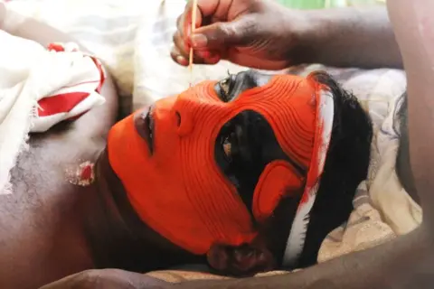 KK Gopalakrishnan Someone laying on their back and having face paint intricately applied by the hand of another person (unseen)