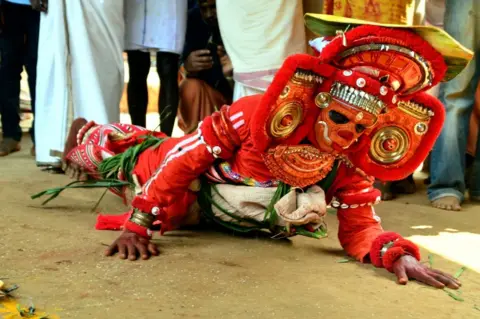 KK Gopalakrishnan A performer lays stomach-down on the floor. They are wearing a red headdress and costume