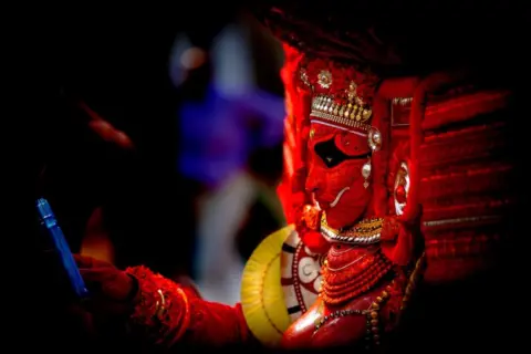 KK Gopalakrishnan A performer in an elaborate red mask and headdress looking at their mobile phone