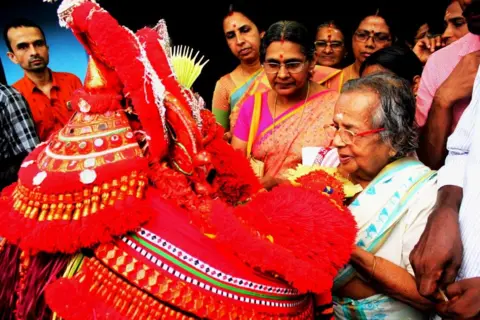 KK Gopalakrishnan An older woman holding a brightly coloured red display, surrounded by other women