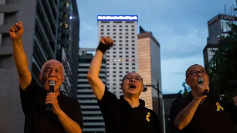 Getty Images Democracy activists Chu You Ming (L), Benny Tai (C) and Chan Kin Man (R), speak on the stage after a march on September 14, 2014 in Hong Kong. They are wearing black t-shirts with golden ribbon pinned to them as they raise their fists in the air. 