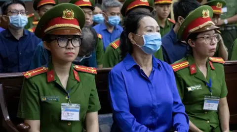 Bloomberg / Getty Images Truong My Lan, chairwoman of Van Thinh Phat Holdings, second left, at the Ho Chi Minh City People's Court in Ho Chi Minh City, Vietnam, on Thursday, Sept. 19, 2024. She is dressed in a blue shirt and has a facemask on. Sat beside her are two women in military uniform.