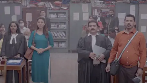 Nila Madhab Panda A still from the film showing a woman wearing a blue kurta standing with her lawyer in a courtroom. To her right, her husband stands wearing an orange shirt and crossbody bag, with his lawyer standing next to them. A thin layer of polluted air hangs in the room