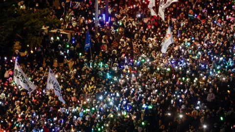 Reuters An aerial view of thousands of people at a protest in south Korea. There are several large banners waving above the crowd, and multiple lights twinkle in the dark.