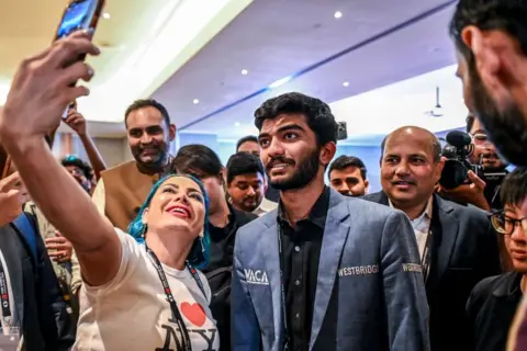 Getty Images Gukesh poses for a selfie with chess fans, he is wearing a blue blazer and black shirt. He is stood next to a woman and is surrounded by people behind.