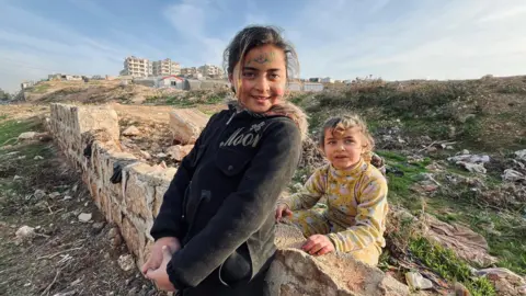 Lee Durant/BBC Two young girls stand leaning against a broken wall in a large rubbish pit at an IDP camp in Idlib