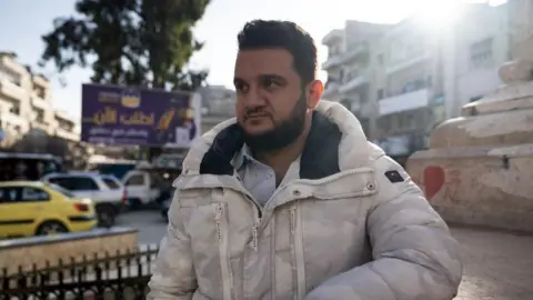 Lee Durant/BBC Young bearded man in white coat stands next to a road in a built-up part of Idlib