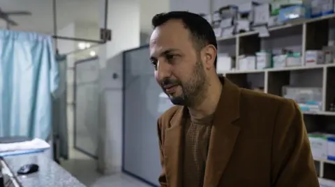 Lee Durant/BBC Doctor stands inside hospital ward with packs of medication in background