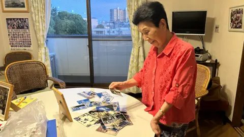 A woman in a pink shirt looks through a pile of black-and-white photographs on a table while standing in front of a window looking out onto a skyline