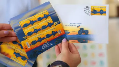 Getty Images Hand holds stamps that read "Good evening, we're from Ukraine"showing a Ukrainian tractor pulling along a Russian tank